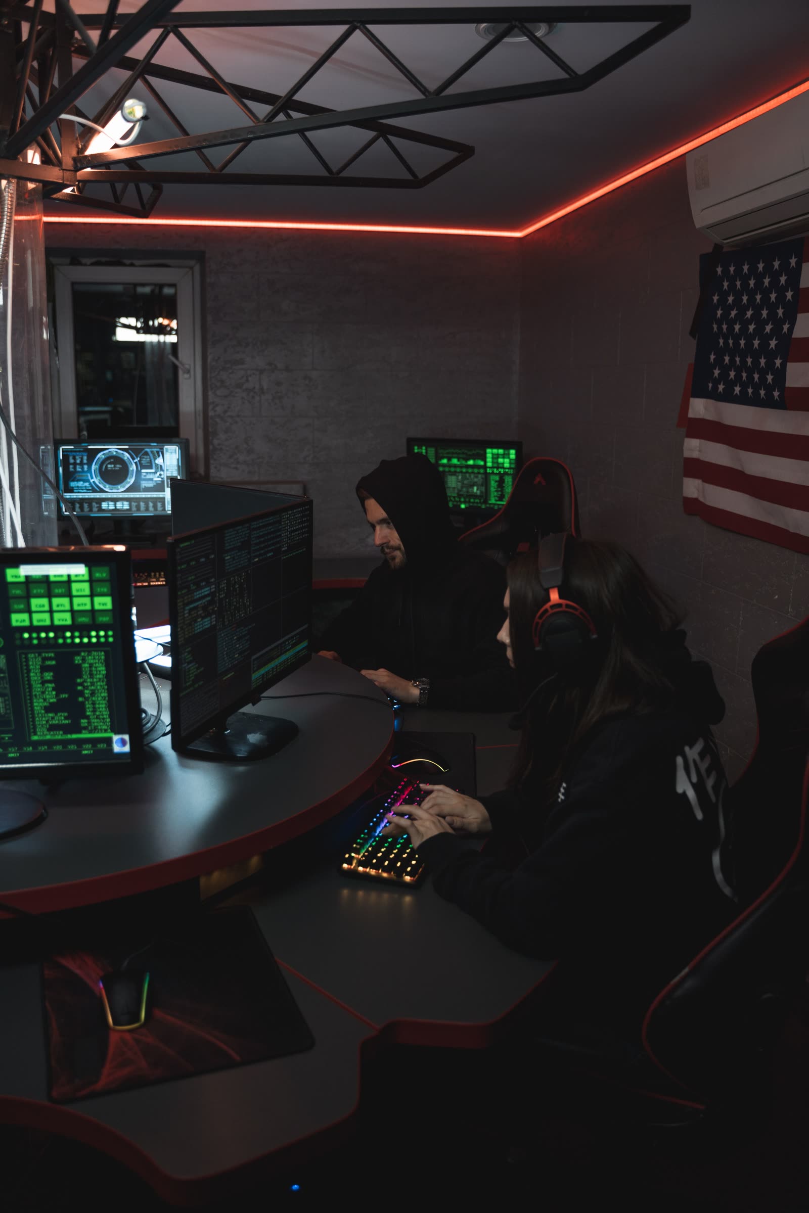 Two players at monitors in a dim red-lit corner room with a flag on the wall