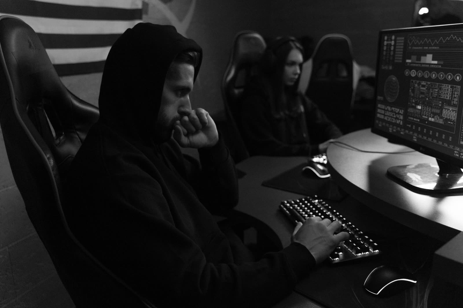Two focused players in a dim, quiet gaming room, one in a hood at a mechanical keyboard