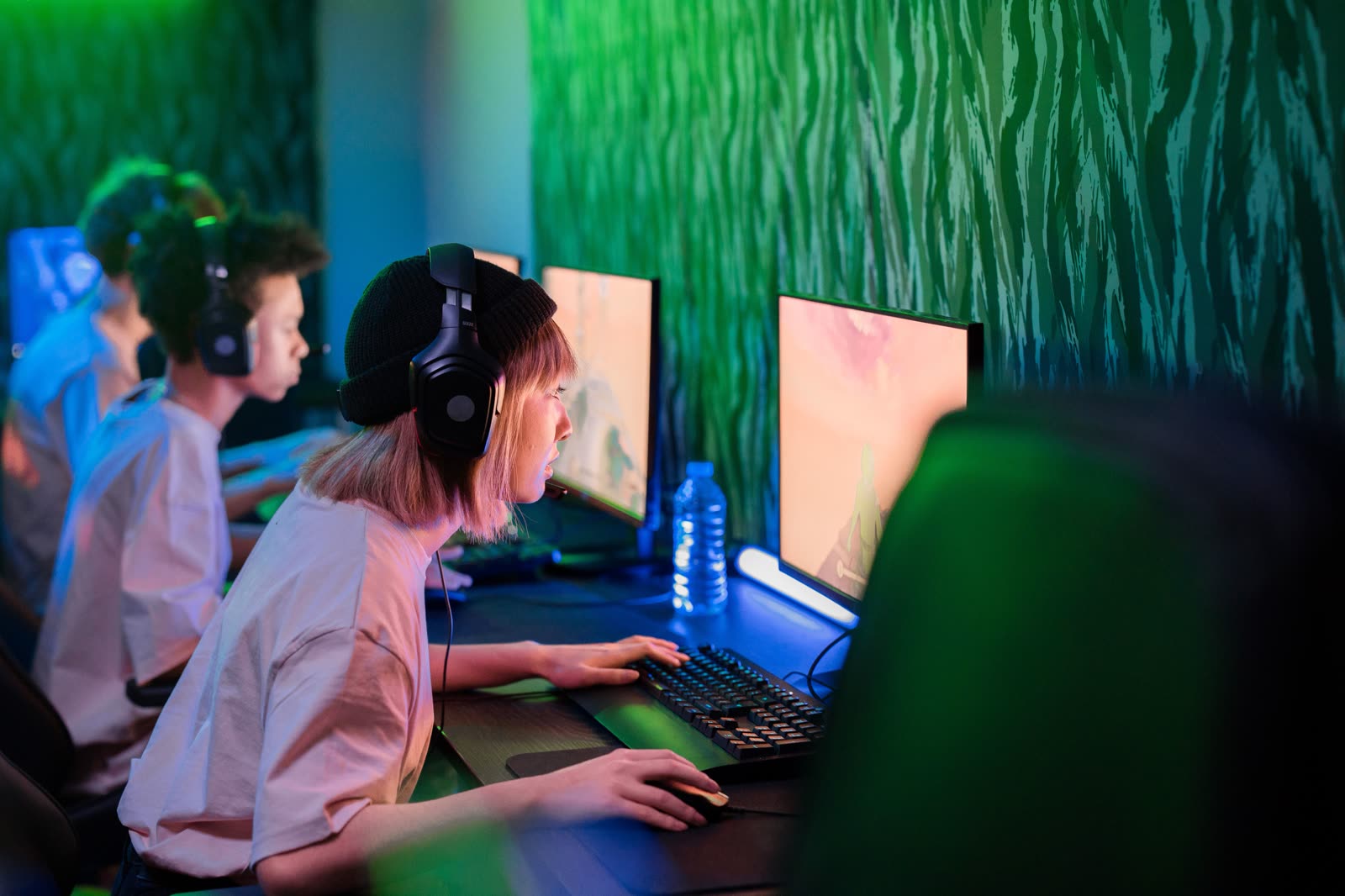 A row of headset-wearing players focused on their monitors along a wall bathed in green neon light